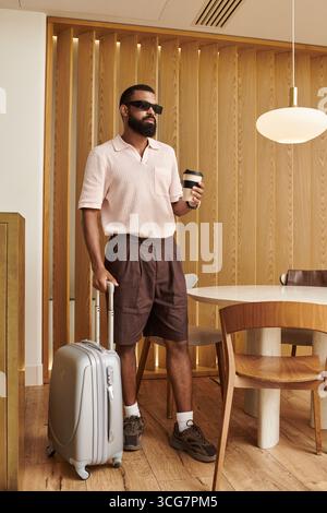 Ein eleganter Afroamerikaner steht in einem modernen Hotelcafé mit Koffer und Kaffee. Stockfoto