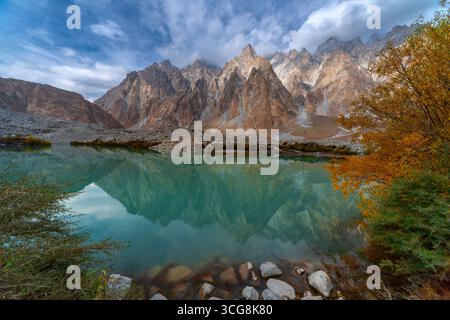 Blick auf zerklüftete Gipfel, die sich in einem ruhigen See spiegeln, umgeben von farbenfrohen Herbstlaub, schaffen einen atemberaubenden Kontrast von Texturen und Tönen in Hunza Nagar, Gilgit Baltistan, Pakistan. Stockfoto