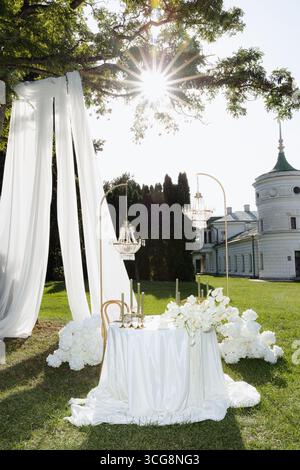 Ein wunderschön gestalteter Tisch mit weißen Vorhängen, floralen Akzenten und Kandelabern in einer üppigen Gartenumgebung unter sanftem Sonnenlicht. Diese Ruhe Stockfoto