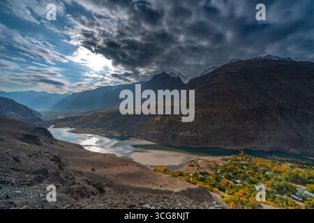 Blick auf das Sonnenlicht, das durch schwere Wolken über einem Tal mit einem gewundenen Fluss und herbstlichen Bäumen bricht, eingerahmt von zerklüfteten Bergen, Yasin Valley, Gilgit Stockfoto