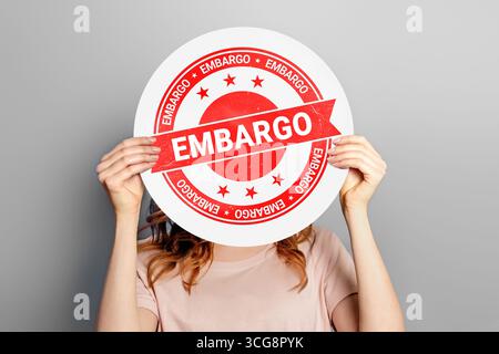 Embargo-Konzept. Frau mit weißem Poster mit Embargostempel isoliert auf grauem Hintergrund im Studio Stockfoto