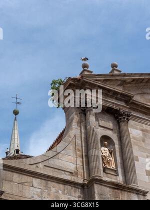 Blick auf ein altes Steingebäude mit einer Statue in einer Nische unter teilweise bewölktem Himmel, ergänzt durch einen Kirchturm mit Kreuz, Alcala de Henares, C. Stockfoto
