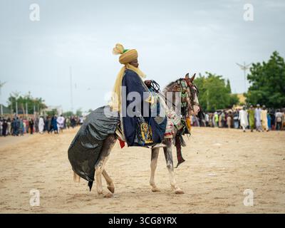 Daura, Nigeria - 15. September 2024: Blick auf eine königliche Figur auf einem Pferd, in lebendiger Kleidung vor dem Hintergrund einer geschäftigen Versammlung und der offenen Landschaft. Stockfoto