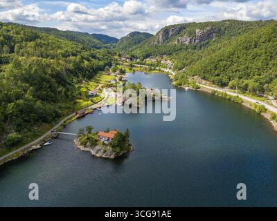 Aus der Vogelperspektive auf einen ruhigen See, der den Himmel reflektiert, umgeben von dichten grünen Wäldern und felsigen Klippen, mit einem Haus auf einer winzigen Insel, Sogne, Agder, Norwegen. Stockfoto