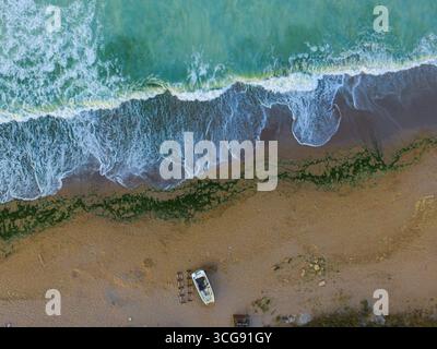 Aus der Vogelperspektive sehen Sie schaumige, türkisfarbene Wellen, die auf die bräunliche Sandküste stürzen, wo ein einsames Boot mit seinem Schatten ruht, Shabla, Bulgarien. Stockfoto