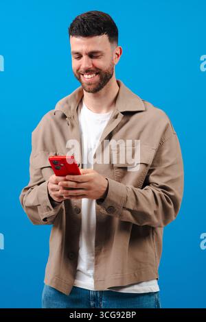 Bärtiger junger Mann, der auf rotem Smartphone schreibt, Jacke auf blauem Hintergrund trägt Stockfoto