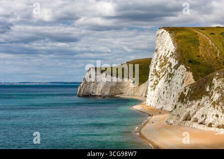 Touristen spazieren entlang des Strandes unterhalb der majestätischen Kreidefelsen in Lulworth, Dorset. Stockfoto