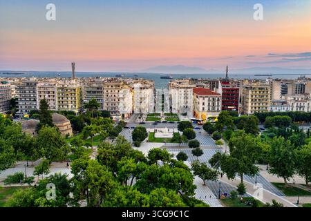 Thessaloniki, Griechenland 3. August 2025. Drohnenansicht des Stadtzentrums bei Sonnenuntergang mit rosa Himmel, städtischer Architektur, Schiffen im Thermaischen Golf und grünem Par Stockfoto