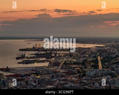 Thessaloniki, Griechenland 3. August 2025 - Panoramablick auf den Hafen von Thessaloniki mit Frachtschiffen, Kränen und der Skyline der Stadt, die während des goldenen Ho beleuchtet wird Stockfoto