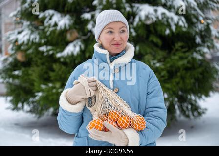 Frau in blauem Wintermantel mit Netztasche im Schnee draußen mit Mandarinen Stockfoto