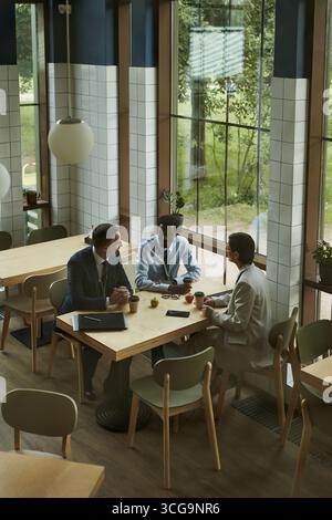 Drei Geschäftsleute mittleren Alters, darunter kaukasische und schwarze Männer und Frauen, sitzen am Tisch in einem modernen Café und diskutieren mit Dokumenten und Kaffeetassen auf dem Tisch über die Arbeit Stockfoto
