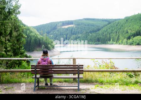 Reisende mit Rucksack, der auf einer Bank sitzt und den friedlichen Blick auf den Bergsee und den Kiefernwald genießt. Konzept von Entspannung, Achtsamkeit und Abenteuer. Stockfoto