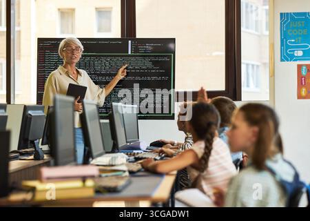 Ältere kaukasische Frau unterrichte eine Gruppe von verschiedenen Kindern, die im Computerunterricht programmieren. Sie stand neben einem großen Monitor, der Programmiercode anzeigt, während die Schüler zuhören und die Hände heben Stockfoto