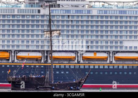 Wismar, Deutschland. August 2025. Die „Wisemara“, eine Nachbildung eines alten Zahnrads vor der Insel Poel, passiert das neue Kreuzfahrtschiff „Disney Adventure“ am ausgerüsteten Kai der Werft von Wismar. Nach rund sieben Jahren Bauzeit wird eines der größten Kreuzfahrtschiffe der Welt in den nächsten Tagen die Hansestadt verlassen. Die US-Reederei Disney Cruise Line übernahm das Schiff vom Insolvenzgut und lässt es von der Meyer Werft in Papenburg, Niedersachsen, bauen. Das Schiff wird für die Disney Group in Südostasien eingesetzt werden Stockfoto