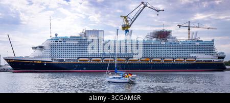 Wismar, Deutschland. August 2025. Ein Segelboot passiert das neue Kreuzfahrtschiff „Disney Adventure“ am ausgerüsteten Kai der Werft in Wismar. Nach rund sieben Jahren Bauzeit wird eines der größten Kreuzfahrtschiffe der Welt in den nächsten Tagen die Hansestadt verlassen. Die US-Reederei Disney Cruise Line übernahm das Schiff vom Insolvenzgut und lässt es von der Meyer Werft in Papenburg, Niedersachsen, bauen. Das Schiff wird ab Ende 2025 für die Disney Group in Südostasien im Einsatz sein. Quelle: Jens Büttner/dpa/Alamy Live News Stockfoto