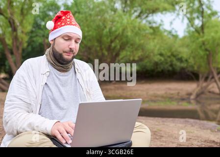 Ein junger hispanischer Mann mit Weihnachtsmütze und Laptop, während er draußen auf dem Boden sitzt, umgeben von der Natur. Stockfoto