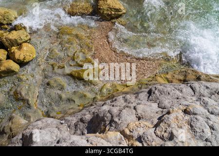 Blick von oben auf die Meereswellen, die über einem strukturierten felsigen und Kiesstrand mit klarem türkisfarbenem Wasser sprudeln. Stockfoto