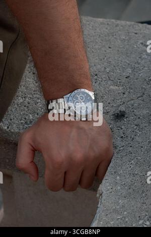 Nahaufnahme der Hand eines Mannes, der auf Beton liegt und eine elegante Armbanduhr aus Stahl mit Metallarmband trägt, modern und stilvoll. Stockfoto