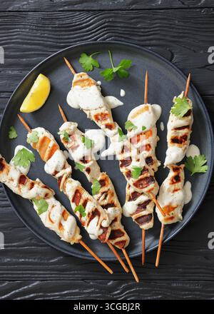 Gegrillte Hähnchenkebabs auf Holzstäben mit Joghurtsauce auf dunkelgrauem Teller auf schwarzem Holztisch, freier Platz, flacher Lay, Nahaufnahme, n Stockfoto