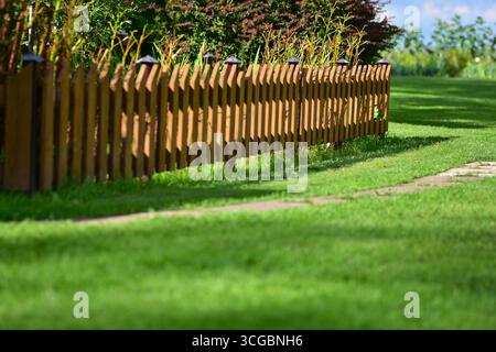 Lange Holzzaun-Linie mit Gartenpflanzen und hellgrünem Gras im Sonnenlicht. Ländliche ländliche Hofgrenze mit natürlichem dekorativem Stil. Stockfoto
