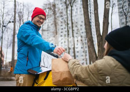 Ein lächelnder Liefermann überreichte einer Kundin eine Papiertüte mit Essen zum Mitnehmen aus seinem Thermalrucksack in einem lebhaften Stadtpark, umgeben von Bäumen und Stadtleben Stockfoto