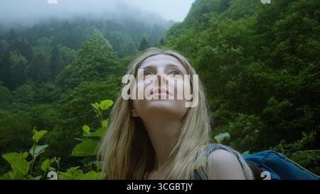 Junge blonde Frau, die bei einem Sommerabenteuer friedlich aufblickt, während sie durch einen üppigen, nebeligen Bergwald wandert. Stockfoto