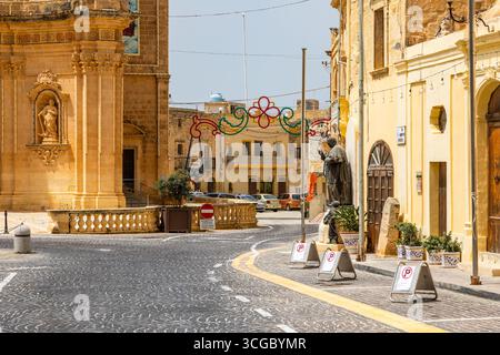 Gozo, Malta – 23. Juni 2021: Straßenszene mit Kirchenfassade, Statue und festlicher Dekoration in einem historischen Stadtzentrum. Stockfoto