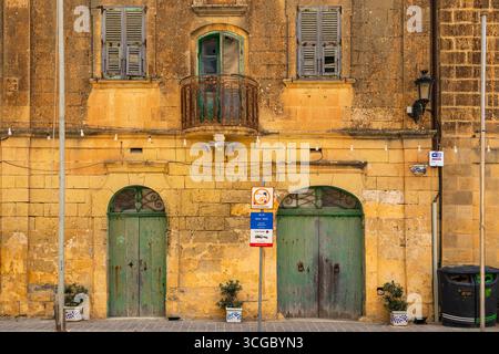 Gozo, Malta – 23. Juni 2021: Verwittertes Kalksteingebäude mit grünen Holztüren, Balkon, Fensterläden und Straßenschildern. Stockfoto