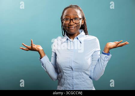 Verwirrte schwarze Frau, die in die Kamera blickt, mit abgezogenen Lippen und zuckenden Schultern. Afroamerikanische Frau spreizt ihre Hände und drückt Unsicherheit mit der Geste „Ich weiß nicht“ aus. Stockfoto