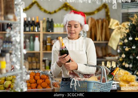 Feale-Kunde im Weihnachtsmann-Hut mit Einkaufswagen kauft eine Flasche Champagner im Supermarkt - bereitet sich auf Weihnachten vor Stockfoto