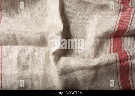 Eine Ansicht von oben nach unten auf ein lässig zerknittertes rustikales beiges Küchentuch mit einem roten Streifen als Hintergrundbild. Stockfoto