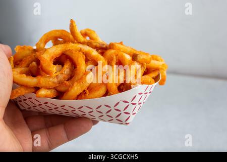 Blick auf eine Hand, die ein Tablett mit gewürzten lockigen Pommes frites hält. Stockfoto