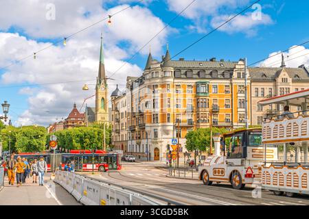 Eine farbenfrohe Straßenszene, während Fußgänger die Djurgardsbron-Brücke entlang laufen, während sie auf die Strandvagen-Straße trifft, mit Touristenzügen, Bussen und dem Uhrturm. Stockfoto
