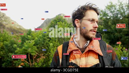 Stehender Mann mit Brille, orangefarbener Jacke, mit schwarzem Rucksack am Hang mit digitalen Ikonen Stockfoto