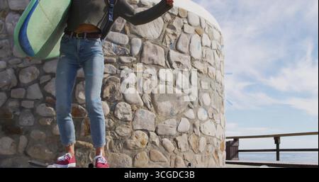 Balancieren Sie Frau, die Skateboard fährt und Surfbrett an der Strandpromenade hält, mit Holzgeländer Stockfoto