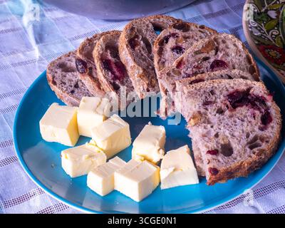 Hausgemachte Obst- und Nussbrotscheiben werden auf einem blauen Teller mit Butterwürfeln angeordnet, um ein einfaches und gesundes Frühstück oder einen Snack zu kreieren Stockfoto
