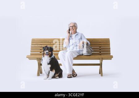 Lächelnde und glückliche ältere Frau, die mit ihrem Hund auf einer Bank sitzt und am Telefon spricht. Stockfoto