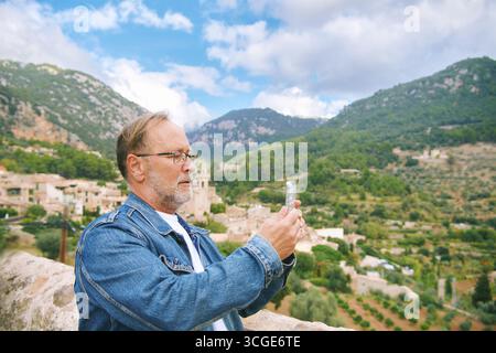 Outdoor-Porträt eines glücklichen reifen Mannes, der einen schönen Tag auf Mallorca, Valdemossa, Spanien, genießt, Fotos oder Videos mit seinem Handy aufnimmt Stockfoto