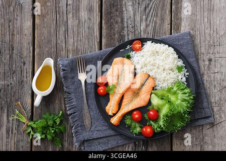 Gebackene Lachssteaks mit Reis, Salat, Tomaten auf einem schwarzen Teller auf rustikalem Holztisch mit Gabel und Serviette, horizontale Ansicht von oben, flache Lage, nicht Stockfoto