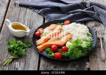 Gebackene Lachssteaks mit Reis, Salat, Tomaten auf einem schwarzen Teller auf rustikalem Holztisch mit Gabel und Serviette, horizontale Ansicht von oben, nicht AI generat Stockfoto