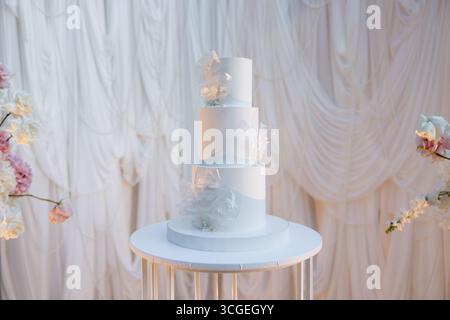 Ein atemberaubender dreistufiger Kuchen steht elegant auf einem Tisch, umgeben von weichem, drapiertem Stoff und Blumenarrangements, die einen romantischen und festlichen Abend einfangen Stockfoto