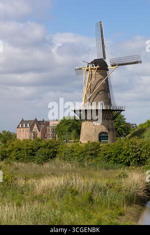 Eine klassische niederländische Windmühle steht hoch zwischen grünen Feldern und einem kleinen Kanal, mit traditionellen Häusern in der Ferne unter hellem Himmel. Stockfoto