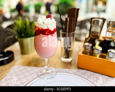 Erdbeermilkshake mit Schlagsahne und Himbeere serviert im Glas am Café-Tisch. Süßes Getränk, Dessert, Erfrischung, Lifestyle und Sommer Stockfoto