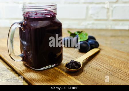 Eine reichhaltige, dunkle Pflaumenmarmelade wird in einem klaren Glasbecher mit Griff auf einem rustikalen Schneidebrett aus Holz gezeigt. Stockfoto