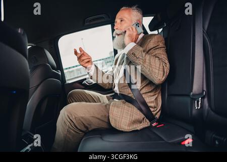 Reifer Geschäftsmann, der auf einem Smartphone auf der Rückbank eines Autos spricht und die Kommunikation in einer stilvollen urbanen Umgebung anregt Stockfoto