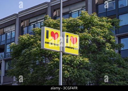 Zwei „I ❤️ Amersfoort“-Schilder mit roten Herzen und dem Namen der Stadt, die an einem Pfosten vor einem Hintergrund grüner Bäume und eines Gebäudes sichtbar sind. Amers Stockfoto