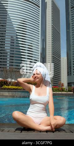 Frau im Badeanzug mit Handtuch auf dem Kopf, die am städtischen Pool sitzt, Stockfoto