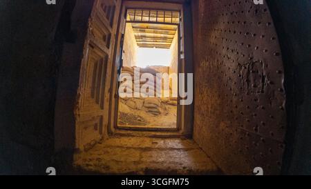Blick durch eine rustikale Tür zu einem sonnendurchfluteten Bereich mit gestapelten Sandsäcken. Ehemalige geheime Militärbasis des IS. Syrien, Palmyra. Stockfoto