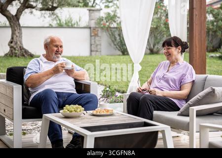 Ältere Männer und Frauen sitzen draußen auf einer Terrasse, lächeln und unterhalten sich, während sie gemeinsam Kaffee und Snacks in einer friedlichen Gartenumgebung genießen. Stockfoto