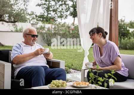 Ältere Männer und Frauen sitzen auf einer Gartenterrasse, lächeln und plaudern beim Kaffeetrinken und genießen gemeinsame Freizeit an einem sonnigen Tag. Stockfoto
